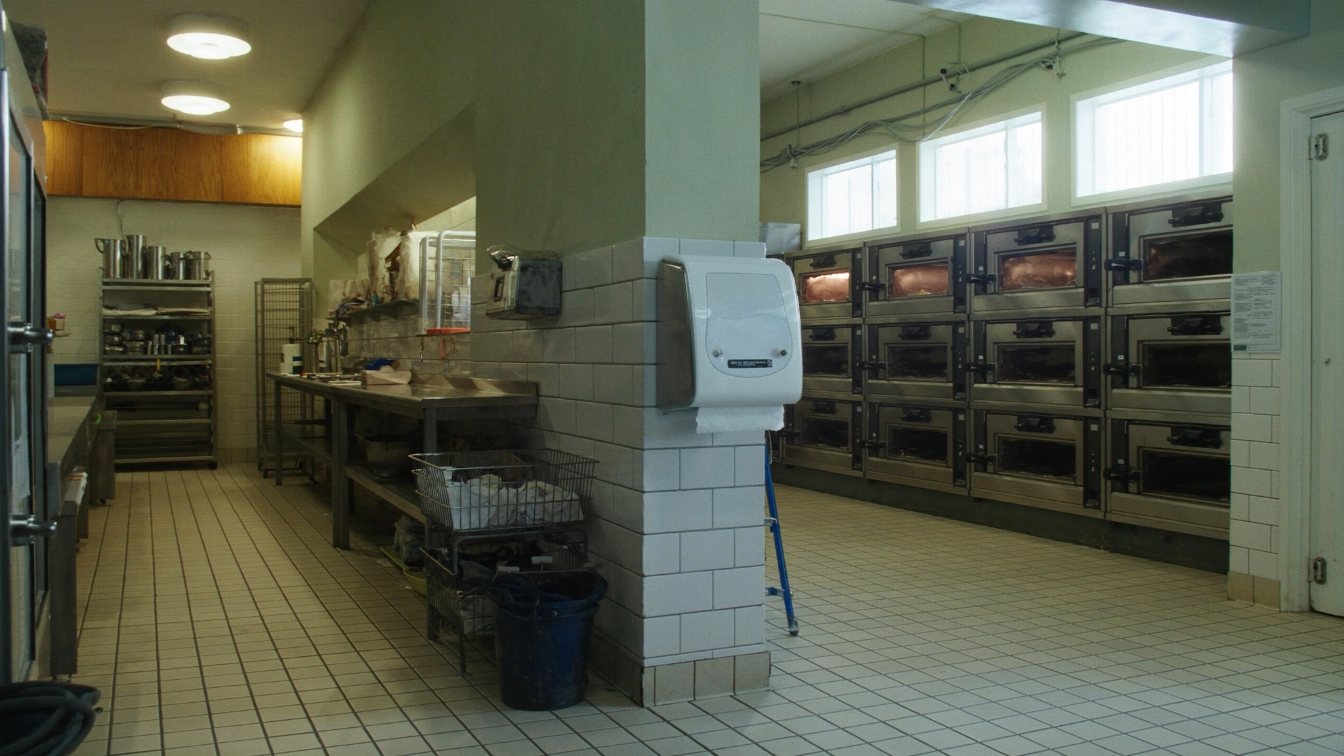 Empty commercial kitchen with stainless steel tables and deck ovens behind a small storefront (illustrative)