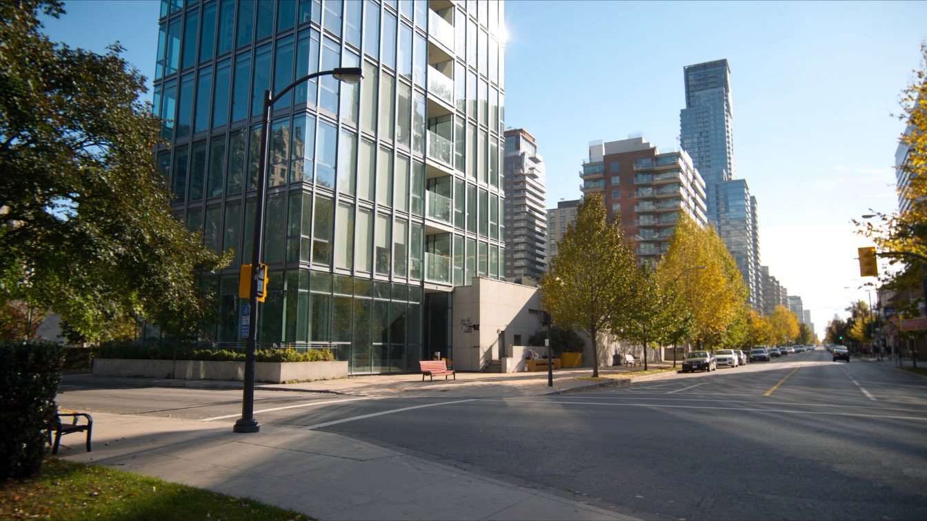 A slim glass condominium tower on a tree-lined downtown Toronto side street with neighbouring towers (illustrative)