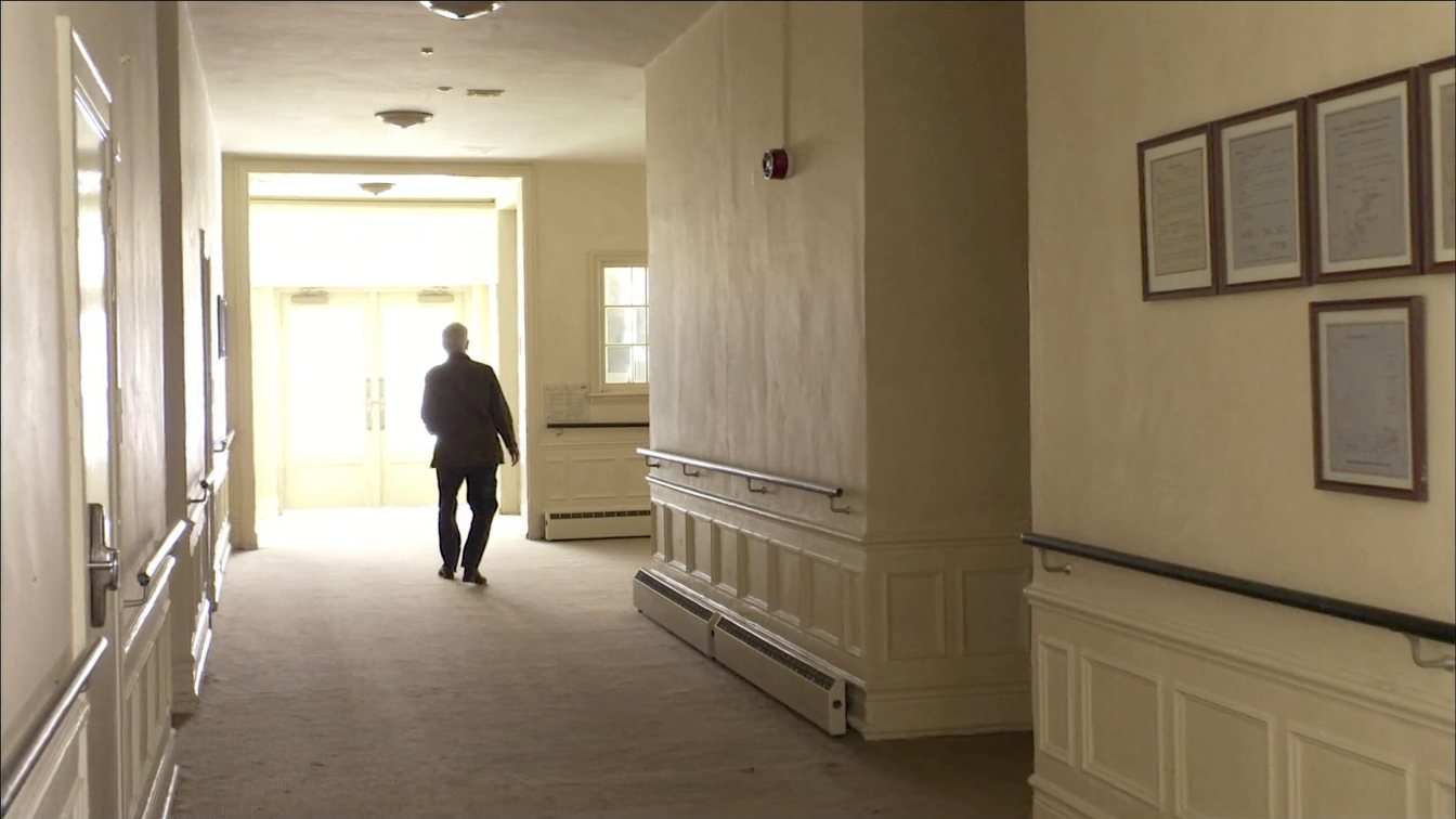 Adult daughter and senior parent walking a retirement residence hallway with handrails, seen from behind (illustrative)