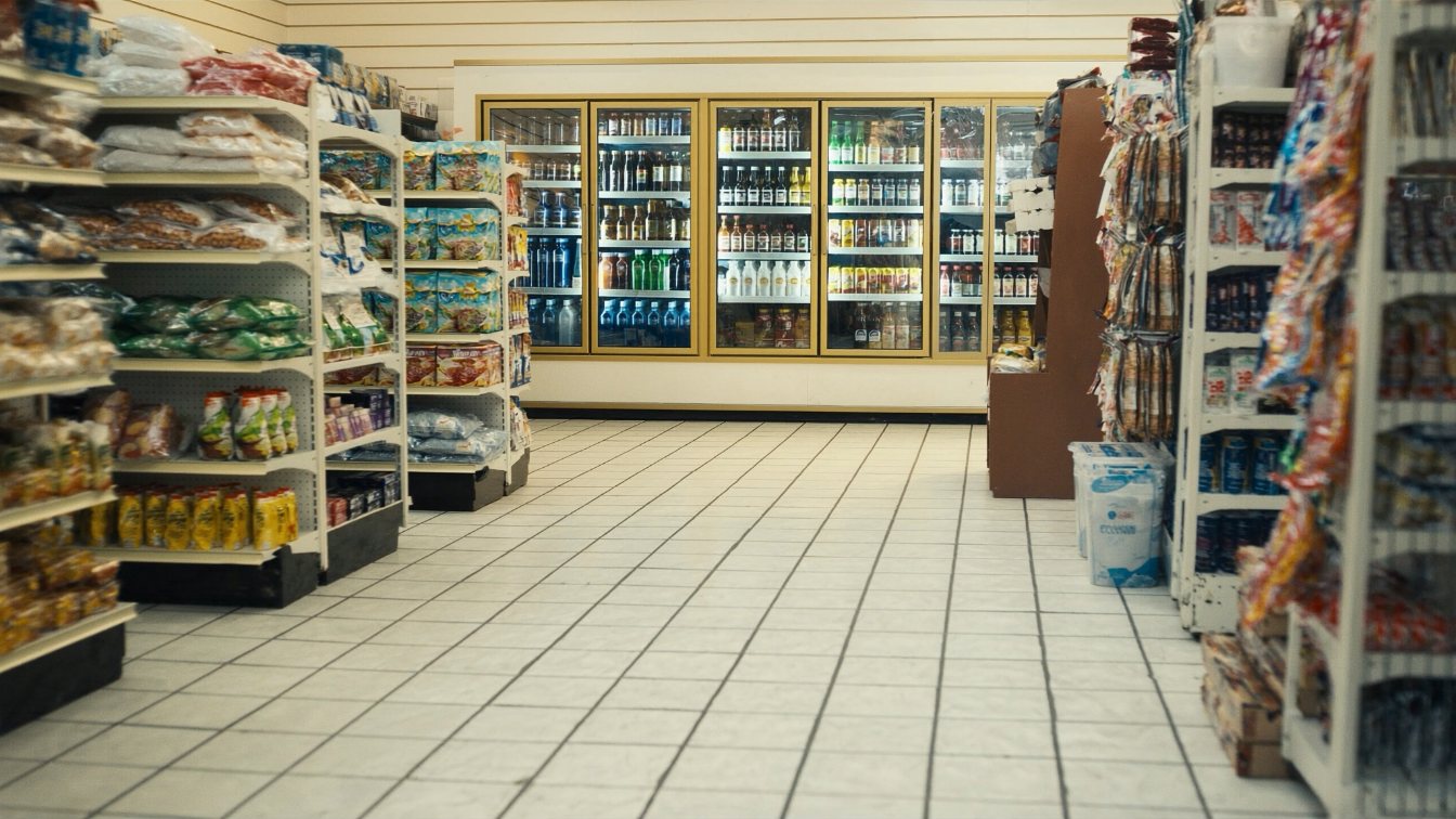 Interior aisles of a small neighbourhood convenience store with grocery shelving and coolers, no people (illustrative)