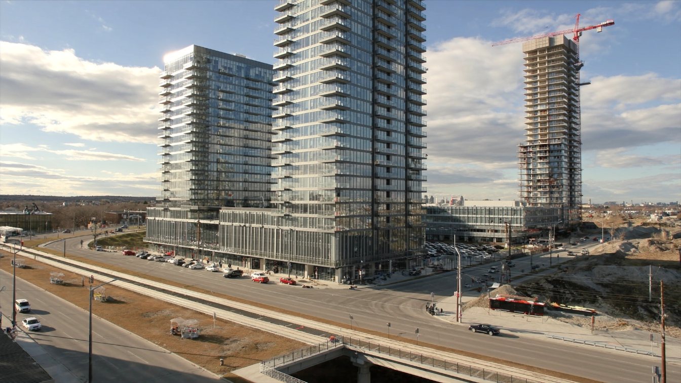 Two completed glass condominium towers beside a third tower under construction on a wide arterial road with light-rail tracks (illustrative)