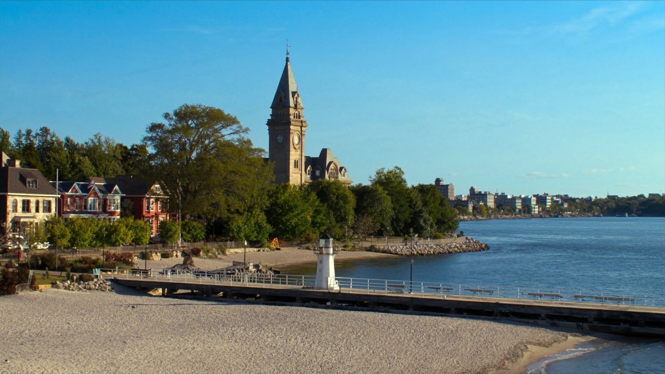 Long white-sand beach with a lighthouse pier and a Victorian town hall tower behind on Lake Ontario in Cobourg (illustrative)