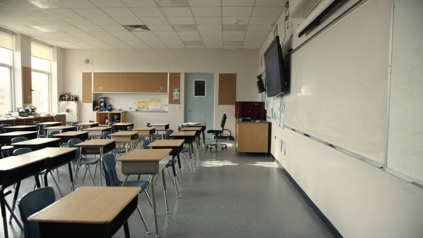 Empty classroom with desks, a whiteboard and daylight from tall windows (illustrative)