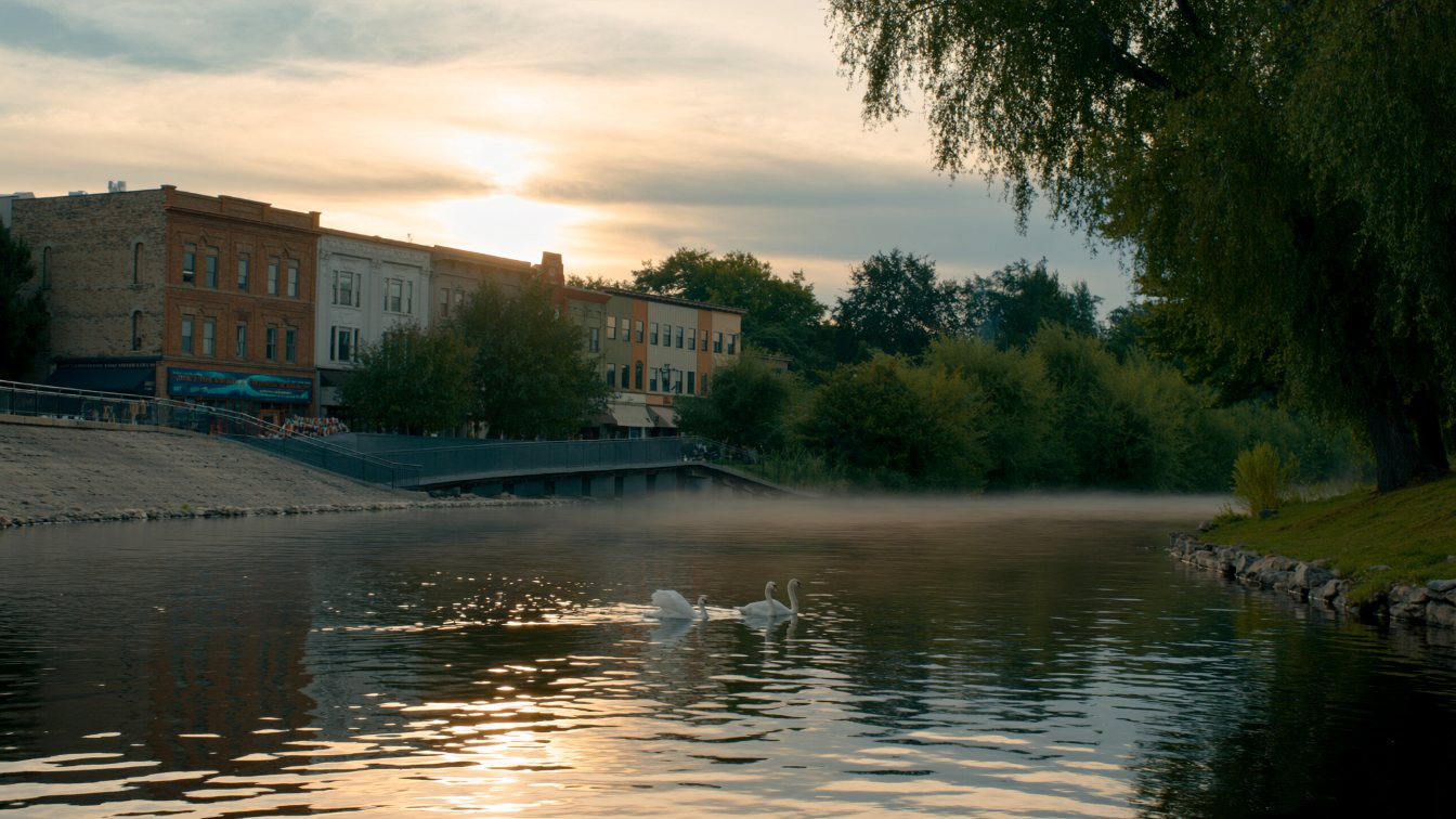 Riverside park with swans and a historic small-town main street beyond, summer evening (illustrative)