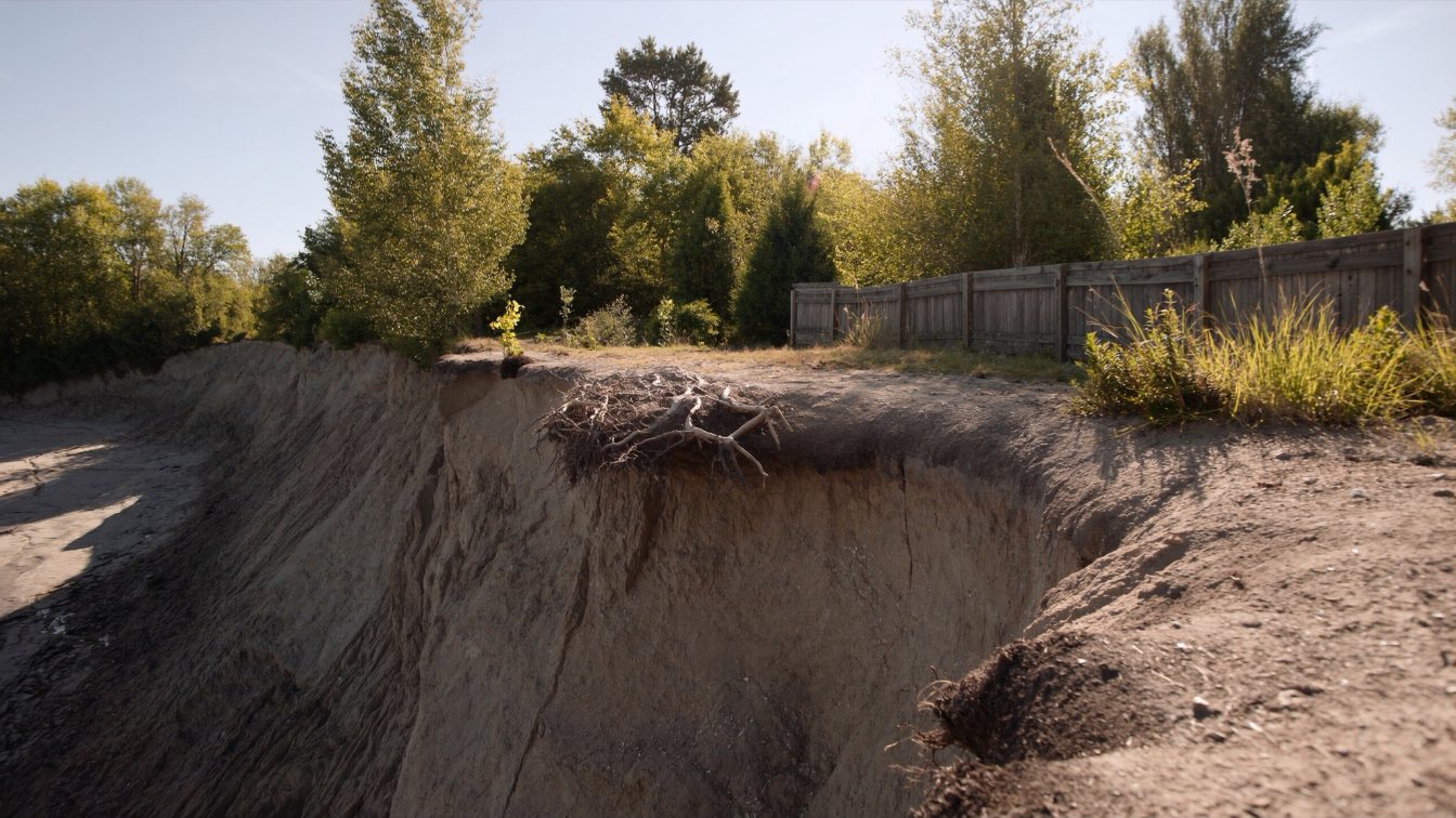 Eroded lakeshore bank with exposed roots and a surveyor's stake near the edge above Lake Ontario (illustrative)