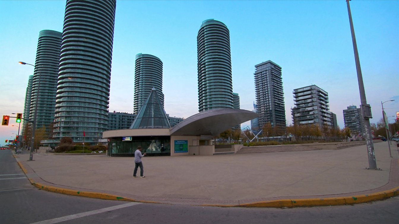 Tall glass condominium towers rising around a subway station plaza in a new suburban downtown at dusk (illustrative)