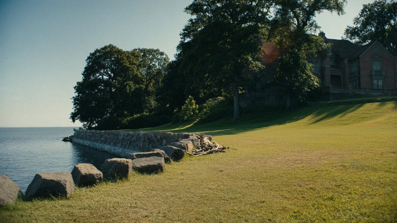 Wide lawn sloping to a stone-protected Lake Ontario shoreline in front of a large older house, late afternoon (illustrative)