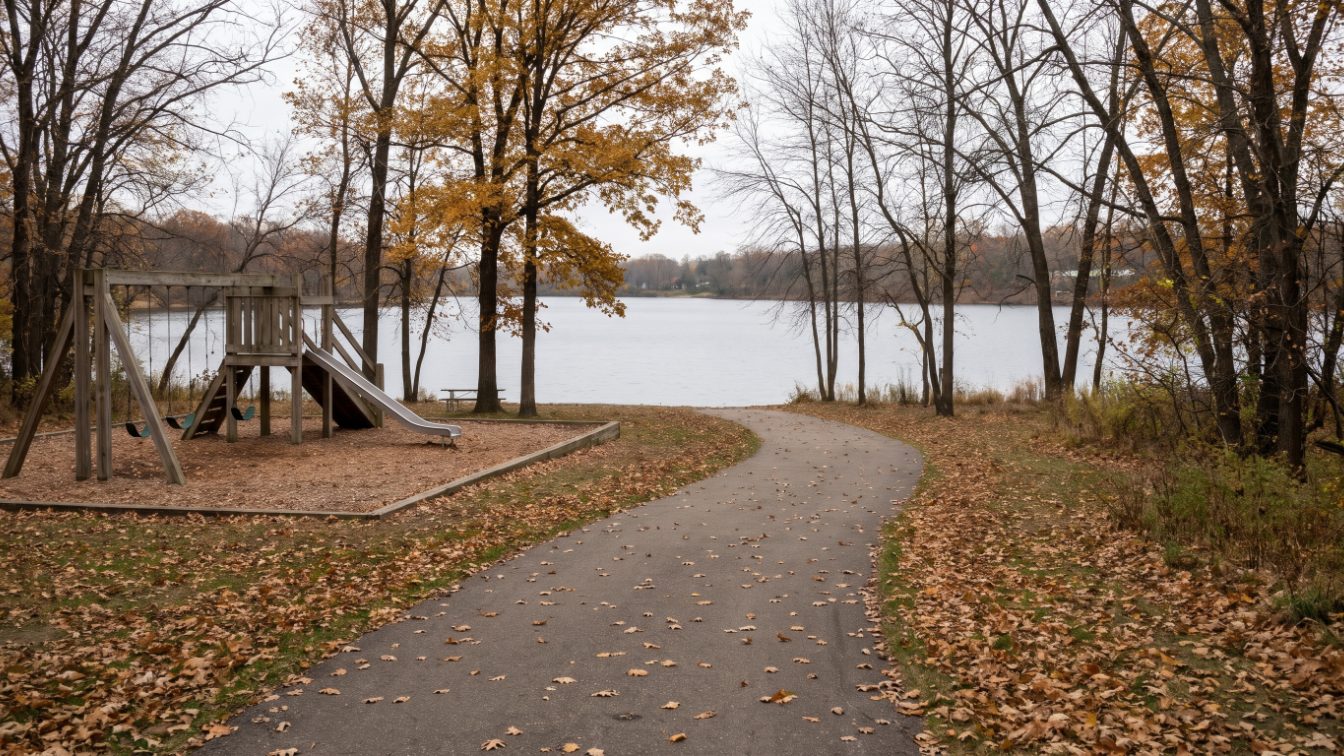Empty lakeside park playground and paved path among autumn trees beside a calm lake (illustrative)