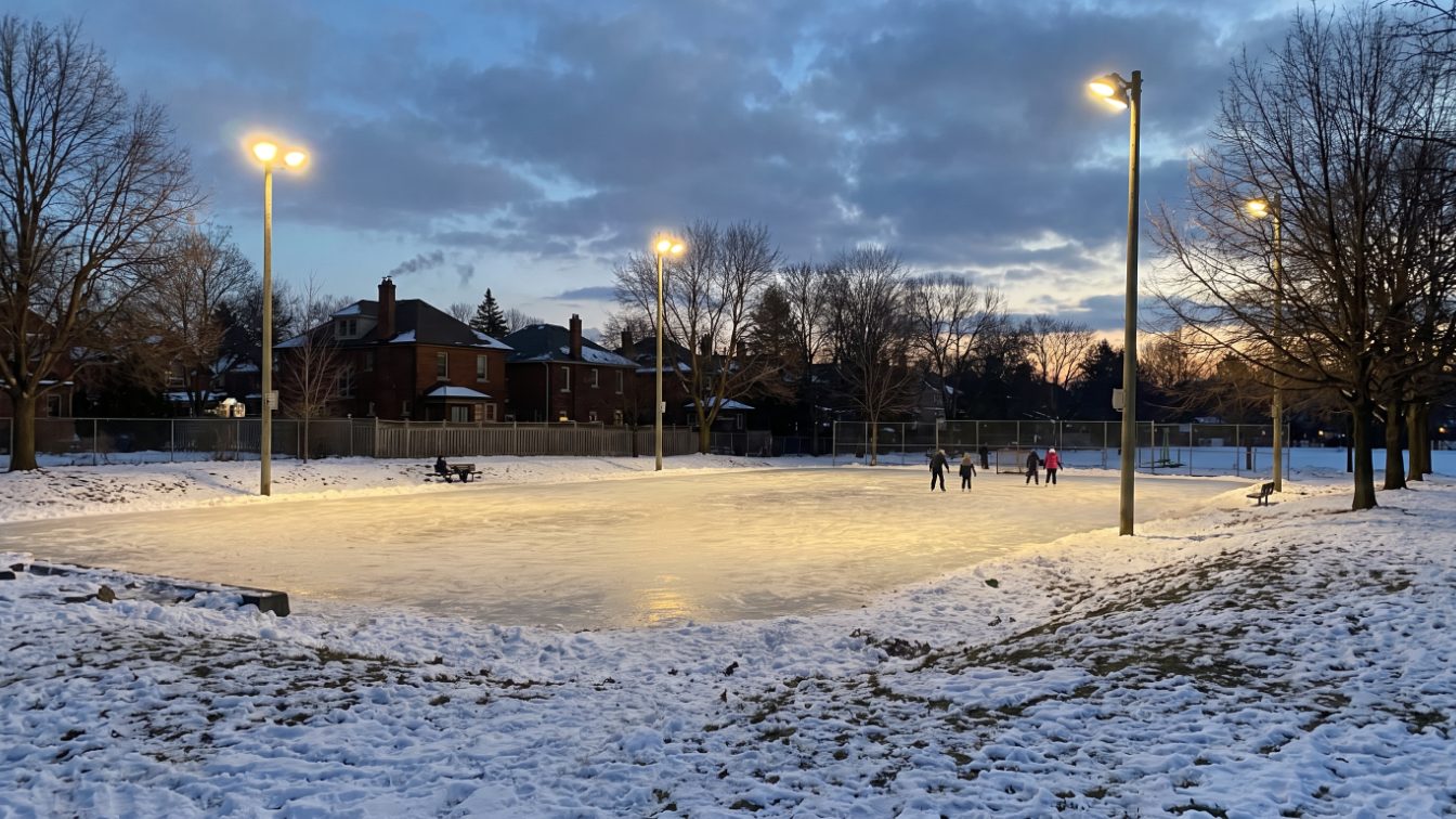 Outdoor neighbourhood ice rink in a snowy park at dusk with distant skaters and houses beyond (illustrative)
