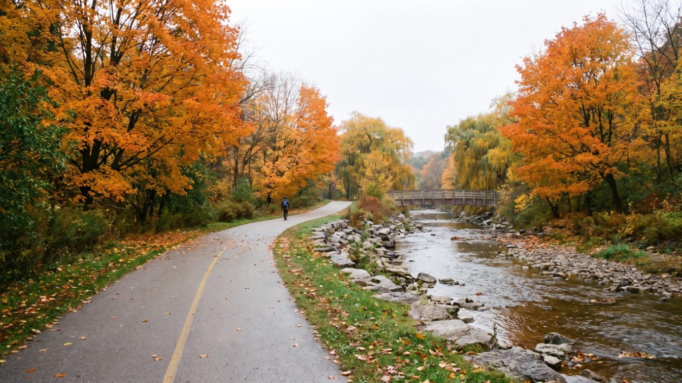 Paved trail through an autumn river valley with a shallow river and distant footbridge (illustrative)